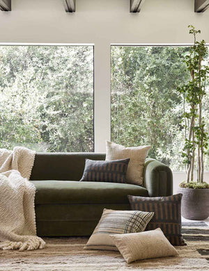 The Kisha natural-toned square throw pillow sits on a green velvet sofa with other throw pillows in a room with large windows