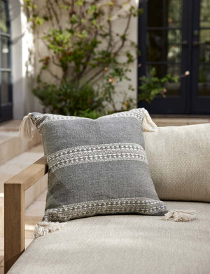 Marchesa agate gray indoor and outdoor square pillow with tasseled corners sits on a natural linen sofa in an outdoor space