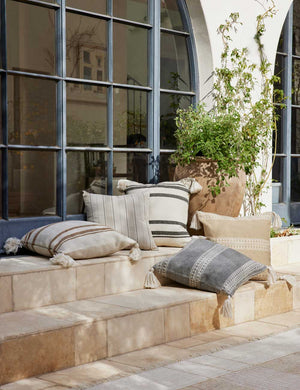 The Fez camel and white indoor and outdoor throw pillow sits on a stone staircase outside with other pillows