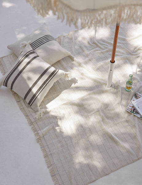 #color::black | The Fez black and white indoor and outdoor throw pillow lays on an ivory outdoor blanket under a fringed beach umbrella