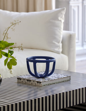 The Moth blue ceramic fruit bowl sits on a book atop a black and white striped coffee table in front of a white linen sofa