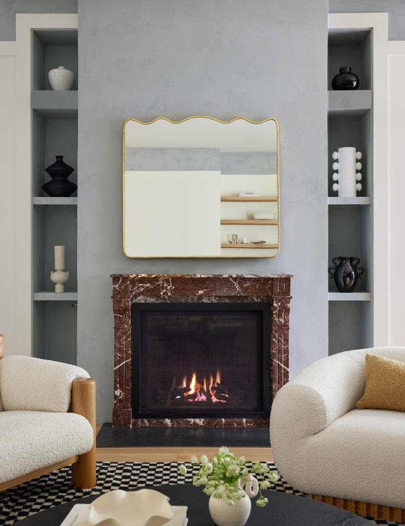| The rook mantel mirror hangs above a burgundy marble fireplace in between two boucle chairs and wall shelves