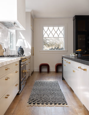 Black and white Checkerboard Rug by Sarah Sherman Samuel in its runner size lays in a kitchen with marble counter tops and golden hardware