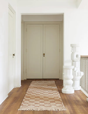 The Irregular beige checkerboard runner rug by Sarah Sherman Samuel sits in a hallway with sculptural pedestals atop a dark hardwood floor.