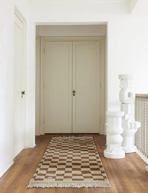 The Irregular ochre checkerboard runner rug by Sarah Sherman Samuel sits in a hallway with sculptural pedestals atop a dark hardwood floor.
