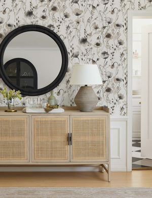 The Scalamandre Lo soft and pastoral brown toned floral wallpaper is in an entryway with a woven light wooden sideboard, a black framed circular mirror, and a lamp with a sculptural base