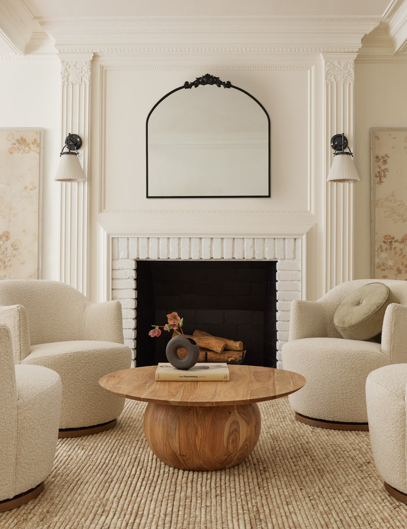 #color::oil-rubbed-bronze | The Tulca arched oil rubbed bronze mirror with flat bottom edge and traditional scroll detailing sits atop a fireplace in a neutral living room with a wooden circular coffee table in the center surrounded by white boucle accent chairs.