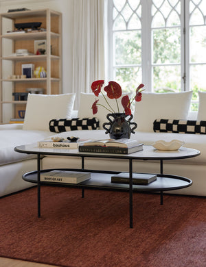 The Ayana Oval Coffee Table sits atop a burgundy rug with books, a black glossy vase, and a white ruffle bowl sitting atop it