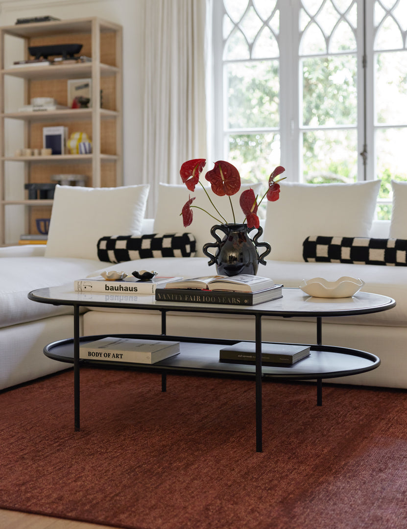 | The Ayana Oval Coffee Table sits atop a burgundy rug with books, a black glossy vase, and a white ruffle bowl sitting atop it