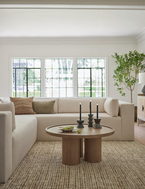 The Delta ash wood round coffee table with three column legs sits in a living room with an ivory modern sectional sofa and a brown hued subtly checked rug.