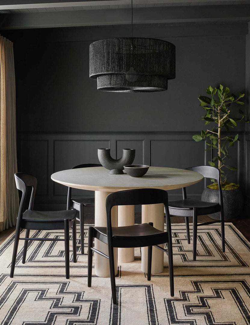 #color::whitewash | Mojave Round Dining Table sits in a room with black accented walls, black dining chairs, and a black and neutral patterned rug