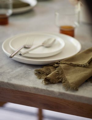 The Set of 4 olive green Essential Cotton Dinner Napkins by Hawkins New York sits on a stone dining table in a dining room with white dinnerware