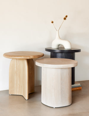The Luna washed-oak wood round side table is next to another wooden side table and the black Luna side table with a white vase