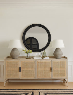 The Philene natural mango wood sideboard with cane doors sits underneath a round black framed mirror with two sculptural gray vases and a white tray sitting on its surface.