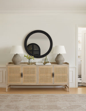 The Yamina taupe indoor and outdoor rug lays in a room with a rattan sideboard and a black round mirror