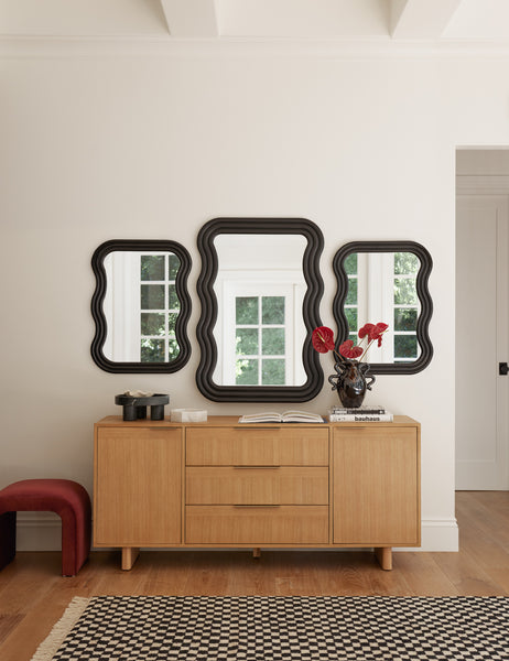 #size::small #size::large | Three Wendolyn wavy thick-framed black wall mirrors hang above a light wood dresser and a black and white checkerboard rug. | The Wendolyn large mirror hangs above a wooden sideboard with the wendolyn small black mirrors hanging to the left and right