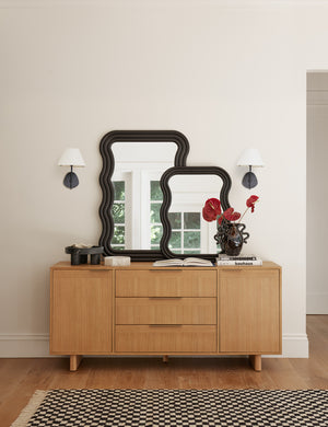 Two Magdalene black single sconces are mounted around two black ripple mirrors and a wooden sideboard