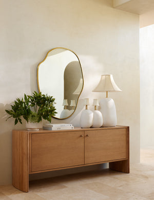 The large puddle mirror hangs on a cream wall over a wooden sideboard that has three sculptural lamps atop it