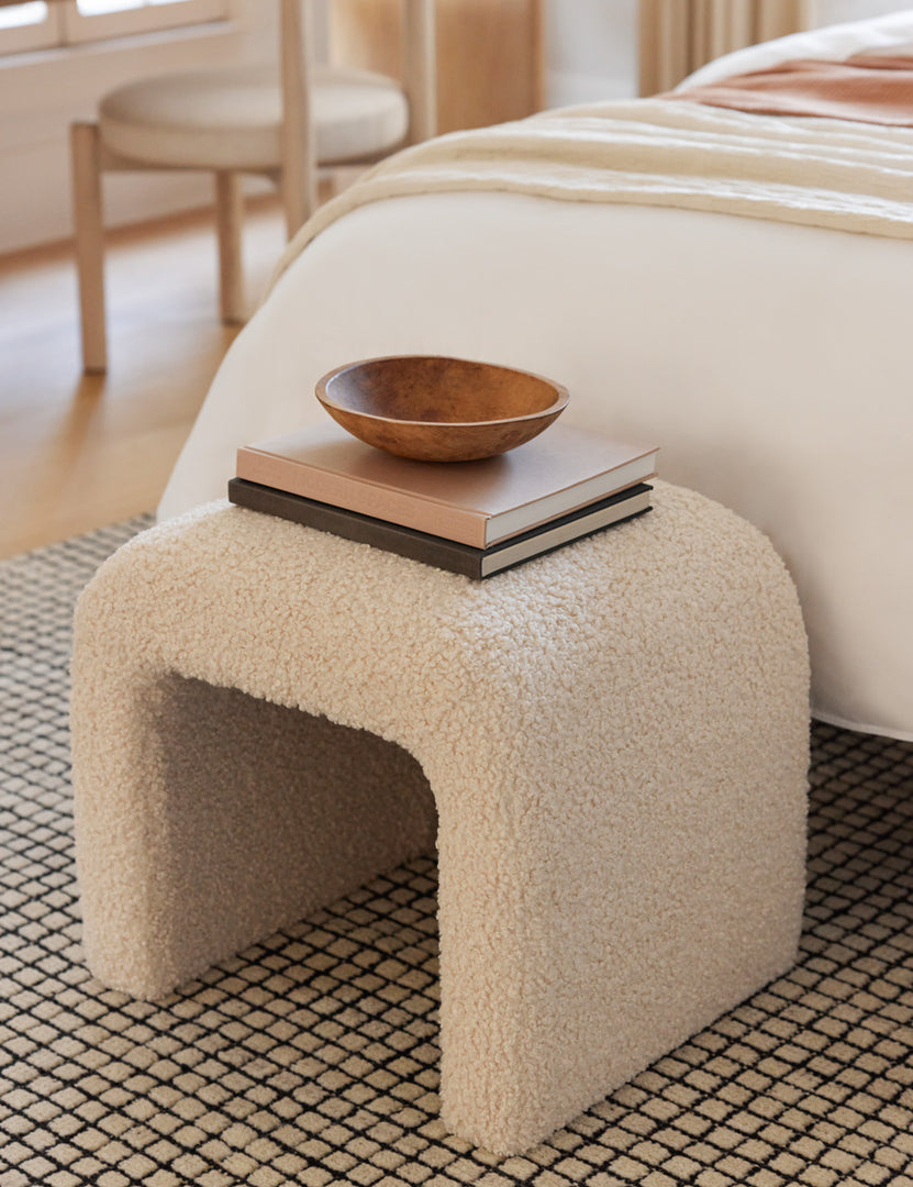 #color::cream | The Tate Cream Boucle stool sits at the end of a bed on a gridded rug with a wooden bowl and a stack of books sitting atop it