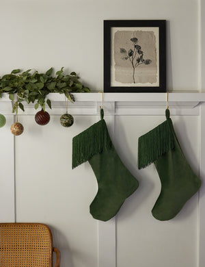 Two Jo Emerald Green Stockings hand from an accented white wall beneath a floral wall art and next to various ornaments and an olive garland