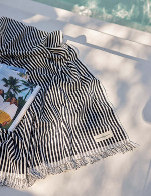 Navy and white striped Beach Towel by Business & Pleasure Co sits by a pool with a book
