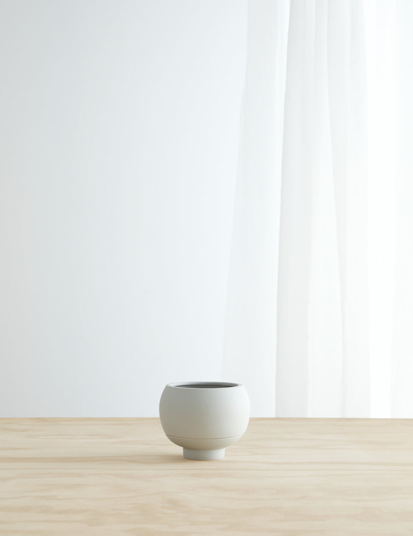 #color::cloud | Sutton Ceramic Self-Watering Planter by Greenery Unlimited in cloud grey sits on a wooden table