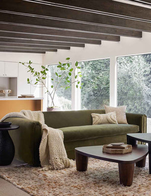 The Eleanor balsam green velvet sofa sits atop a plush rug behind nested coffee tables with an ivory throw atop it