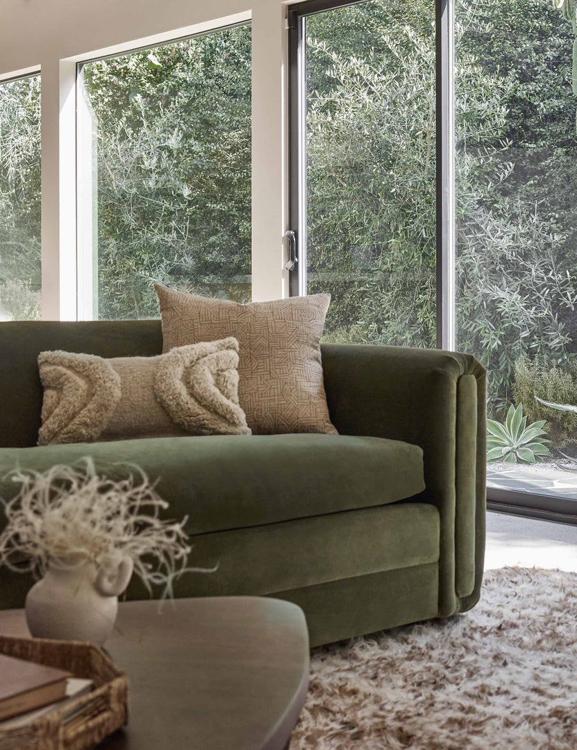#color::natural #size::lumbar | The arches ivory lumbar pillow sits atop a green velvet sofa in a room with floor to ceiling windows