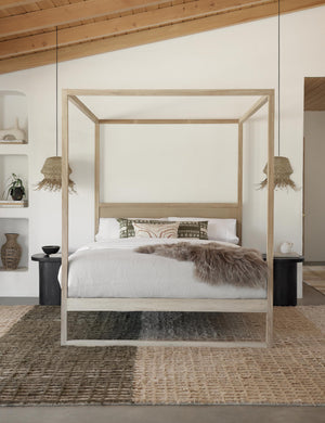 The Kiery light wood canopy bed sits in a bedroom with jute pendant chandeliers, a sheepskin bed cover, and a textured color-blocked rug.