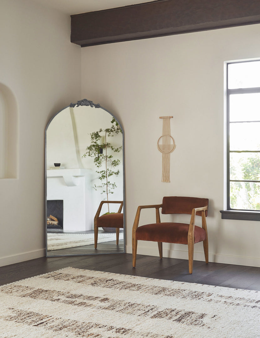 #color::silver | The Tulca silver curved standing mirror with flat bottom edge and traditional scroll detailing sits in the corner of a living room atop a black hardwood floor with a brown velvet accent chair and white and brown patterned rug in front of it.