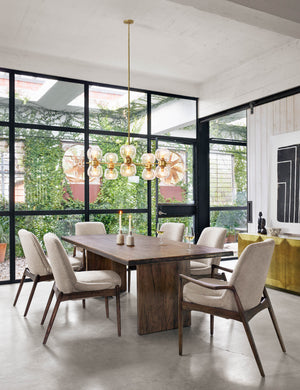 The Ashbie Dining Table sits in a bright dining room surrounded by six ivory dining chairs under a globe chandelier