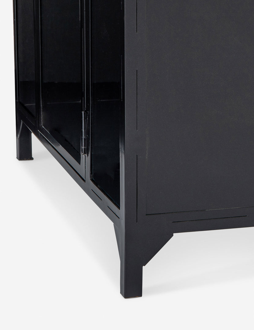| Close-up of the bottom right corner of the Cressida matte black iron curio cabinet with glass paneled doors and brass hardware
