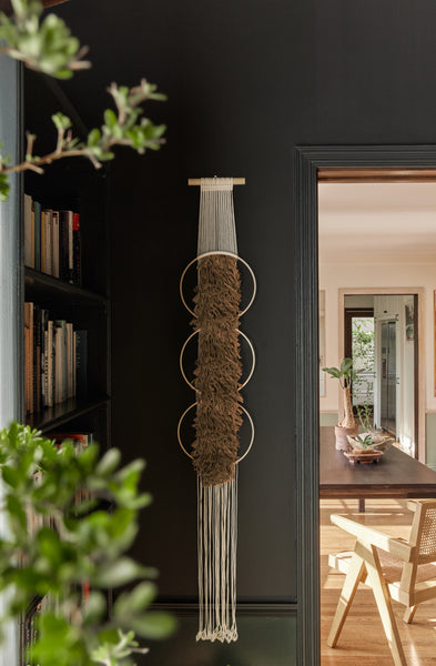 #showall::39842068660323 | The Studio Nom Lwazi light brown woven Wall Hanging by nom hangs in a black painted room next to a bookshelf and a dining room in the background