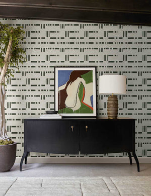 The Artemis green and white dash-and-bar motif patterned wallpaper is in a room with a black wooden console table, an abstract colorful wall art, and a brown ribbed lamp