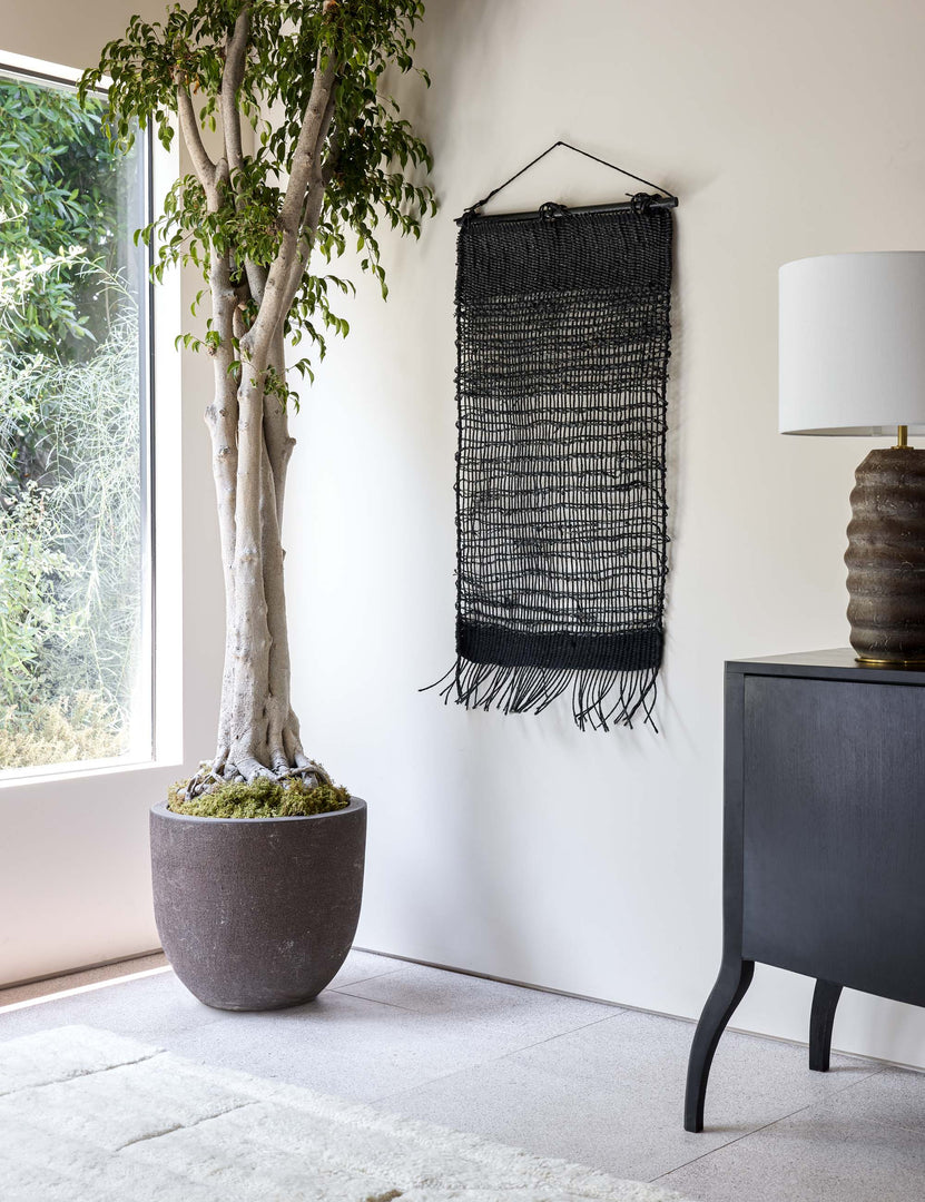 #color::black | The Nyana natural wall hanging hangs in between a large window and a black sideboard