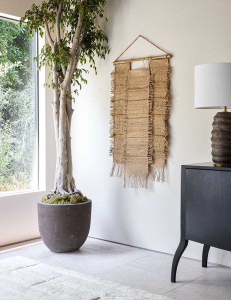 #size::small | The Ukiah woven natural jute and hemp Wall Hanging hangs in a bright room with a ribbed lamp, a black wooden side table, and a plush white rug