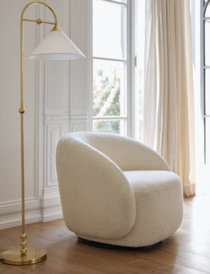 The tauri cream boucle swivel chair sits in a bright room next to a brass floor lamp