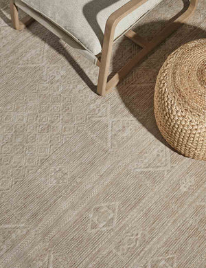 Close-up view of the Yamina taupe indoor and outdoor rug laying under a woven ottoman and a wood-framed chair