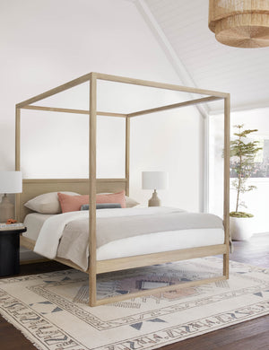 The Kiery light wood canopy bed sits in a bright bedroom with a sloped ceiling, neutral colored bedding, and a multicolored patterned rug.