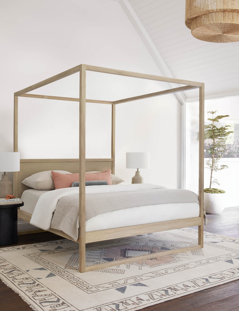 #size::queen #size::king | The Kiery light wood canopy bed sits in a bright bedroom with a sloped ceiling, neutral colored bedding, and a multicolored patterned rug.