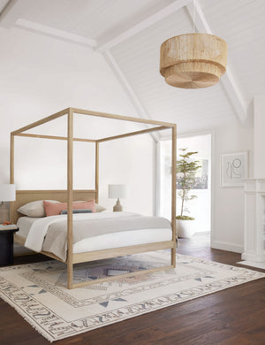 The Sayan natural jute-wrapped pendant light sits in a bedroom with a sloped ceiling, a natural wood canopy bed, and a multicolored geometric rug.