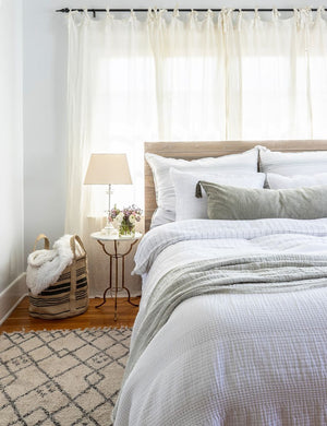 The Blake white and ocean linen duvet by pom pom at home lays atop a wooden framed bed in a bright bedroom with a plush patterned rug and sheer white curtains