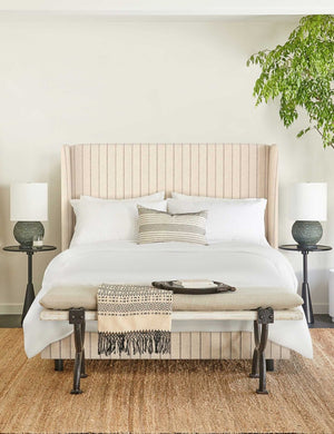 The Adara natural stripe linen upholstered bed sits in a bright bedroom in between two black sculptural nightstands with a cushioned wood bench with metal legs at the end atop a natural jute rug.
