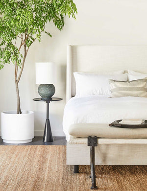 The Adara talc linen upholstered bed sits in a bright bedroom with a black sculptural night stand, a wooden bench with metal legs, all sitting atop a jute natural rug.