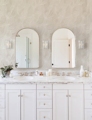 Alina neutral Wallpaper with smooth ripple pattern is in a bathroom with white countertops and cabinetry and two arched mirrors