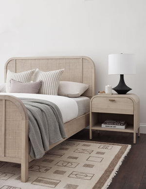 The jillian rug lays in a bedroom under a whitewashed woven framed bed and a whitewashed rounded nightstand