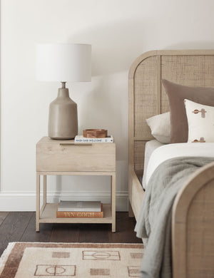 Dana Natural Wood Nightstand sits to the right of a bed with a neutral woven frame that sits atop a brown and cream patterned rug