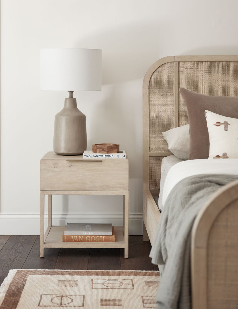 #color::natural | Dana Natural Wood Nightstand sits to the right of a bed with a neutral woven frame that sits atop a brown and cream patterned rug