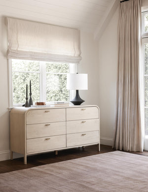 The Brooke whitewashed oak 6-drawer rounded dresser with iron drawer pulls sits against a window with a black lamp and two dark tapered candles on it.