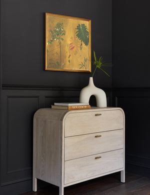 The Brooke 3-drawer white-washed oak dresser sits underneath a yellow floral painting with a stack of books and sculptural vase